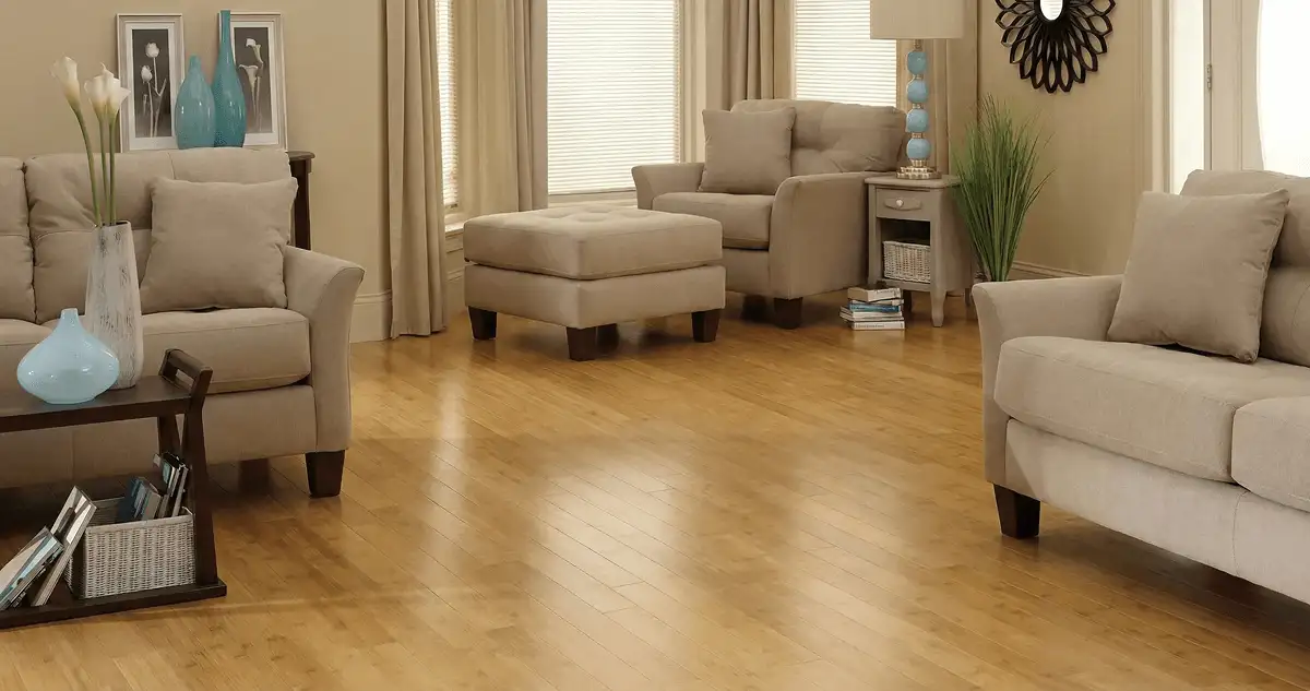 A living room with professionally installed bamboo flooring, beige sofas, a matching ottoman, a side table with a blue lamp, vases, framed pictures, and large windows with blinds and curtains in Chandler, AZ
